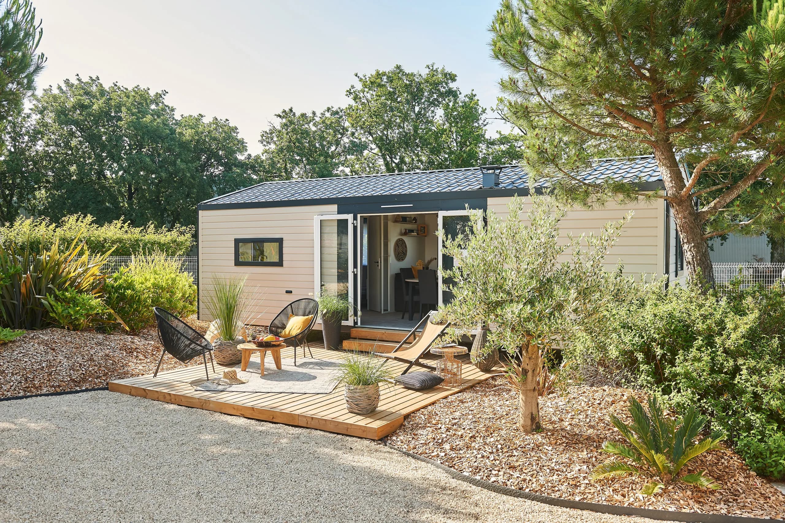 Un mobil-home et sa terrasse en bois baignés de soleil, au cœur d’un jardin arboré
